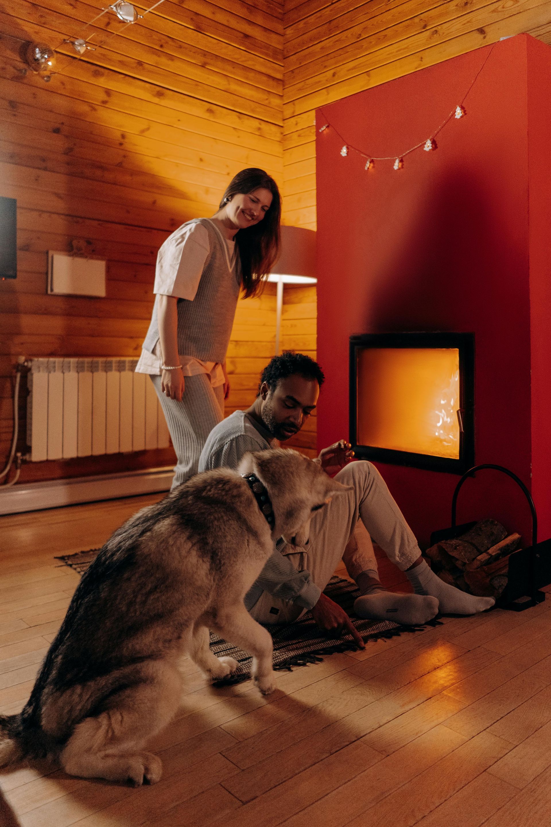 Couple and dog in cozy room with fireplace. Woman smiles at man sitting on floor with dog. Red wall, wooden paneling.