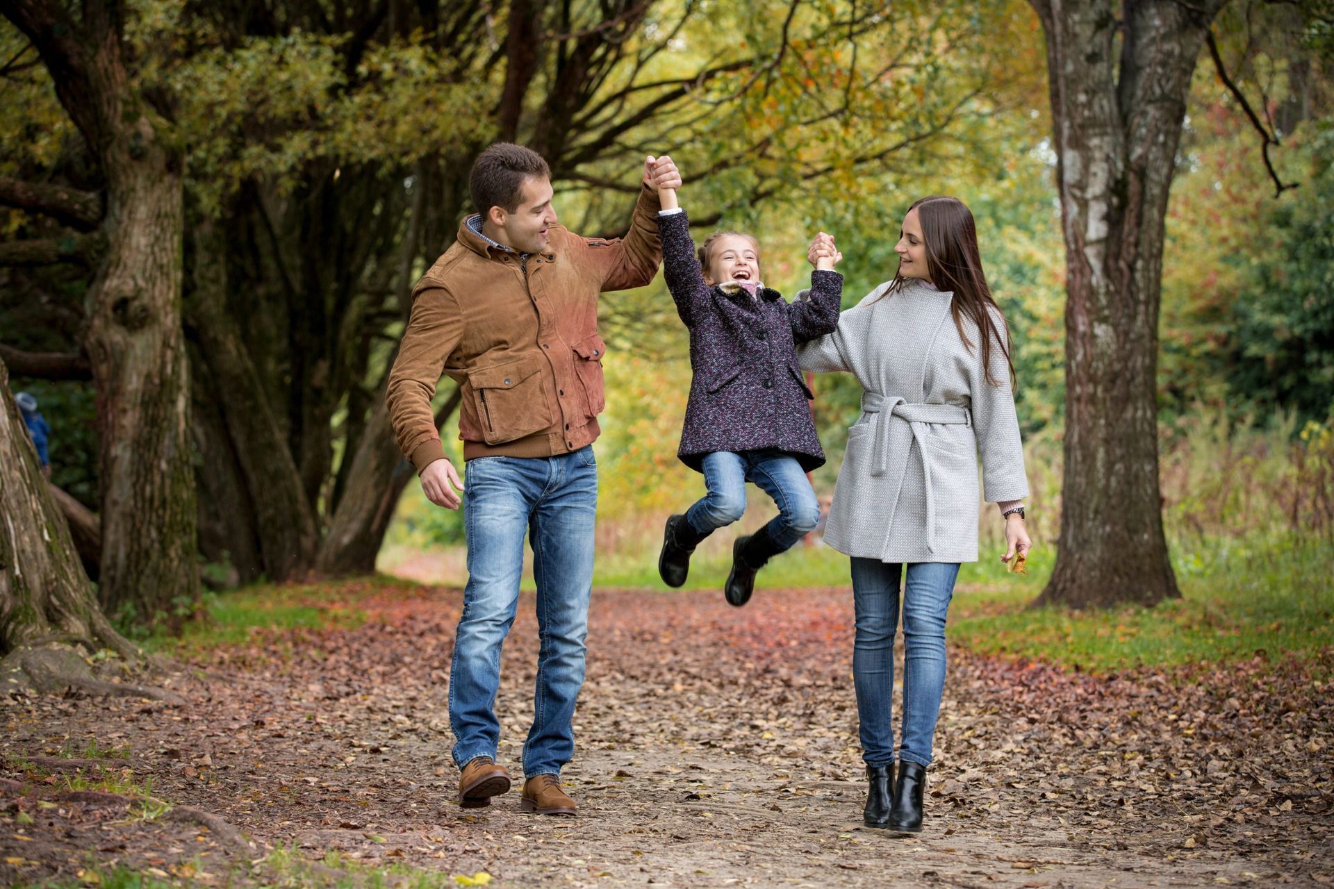 Family in autumn park, father swinging child while mother watches.