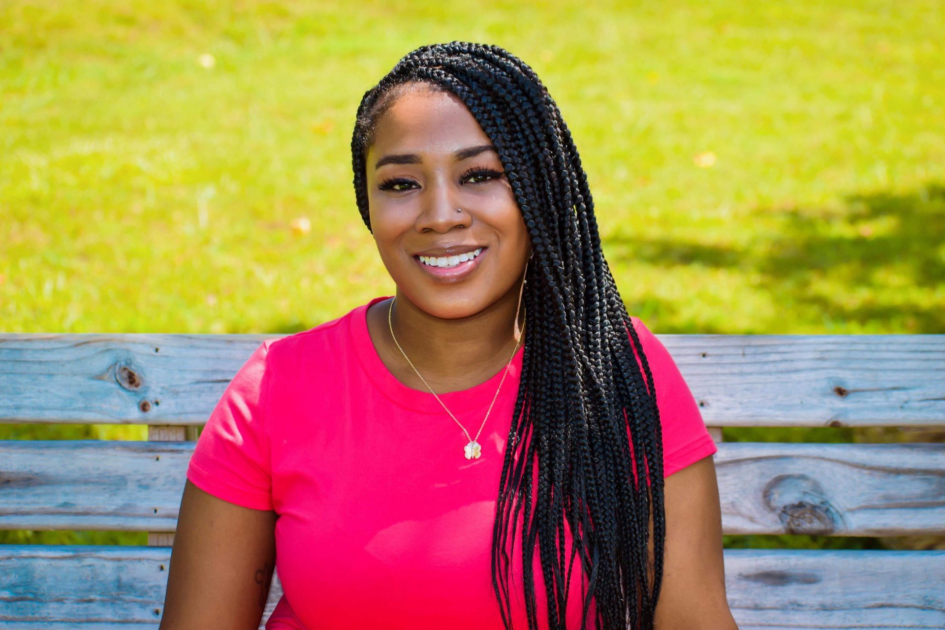 Smiling woman in a pink shirt sitting on a bench in front of a grassy background