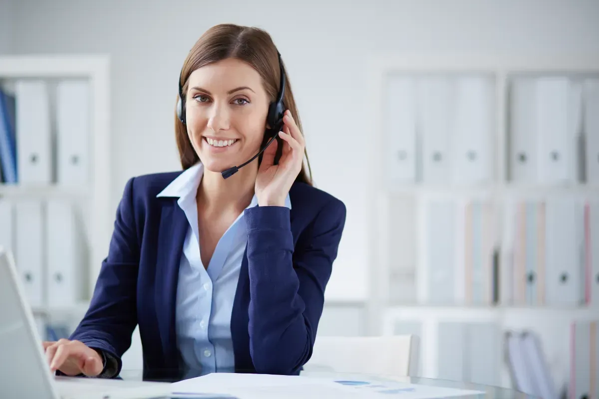 Woman in business attire, wearing headset, smiling at the camera while working at a desk with a laptop.