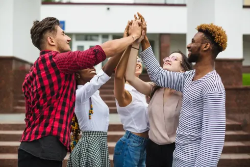 Group of diverse people in front of a building, hands stacked in a high five, smiling in celebration.