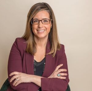 Woman with glasses, arms crossed, smiling in a burgundy blazer, neutral background.