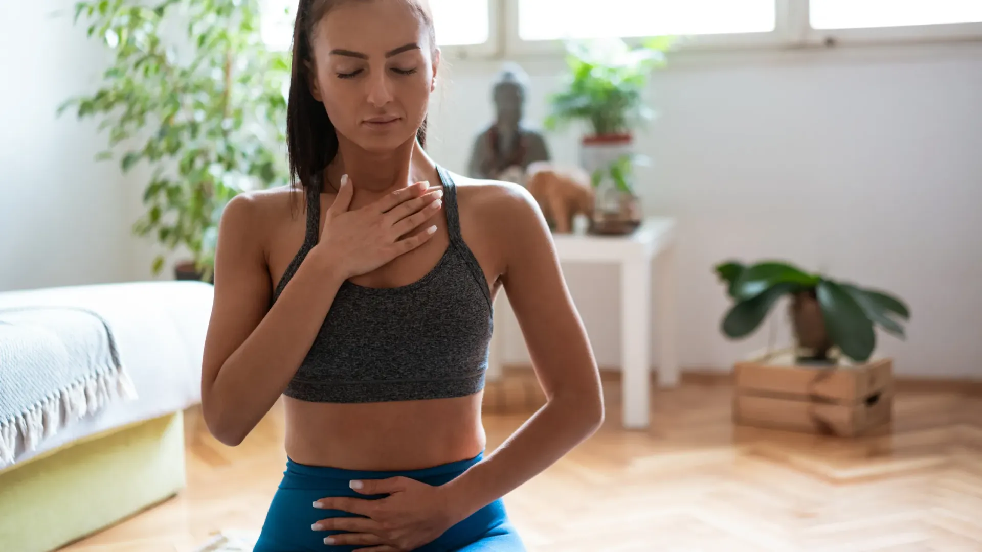 Woman in athletic wear meditating, hands on chest and stomach, eyes closed indoors.