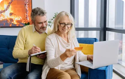 A senior couple on a blue sofa, woman holding a credit card, using a laptop, man writing in a notebook.