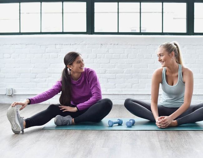 Two women in workout clothes on a mat, stretching and smiling in a bright gym.