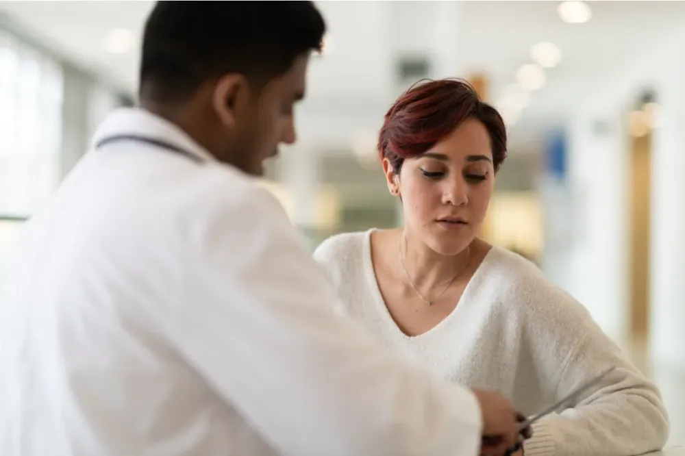 Doctor in white coat consults with a woman; they are indoors in a bright hall.