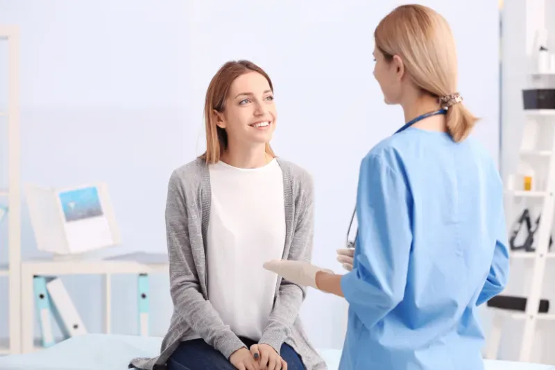Woman in a clinic smiles at a doctor in blue scrubs; light-colored room.