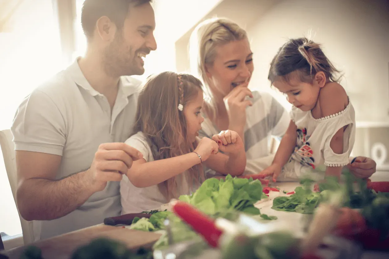 Family of four preparing food in a kitchen; smiling, light-filled scene.