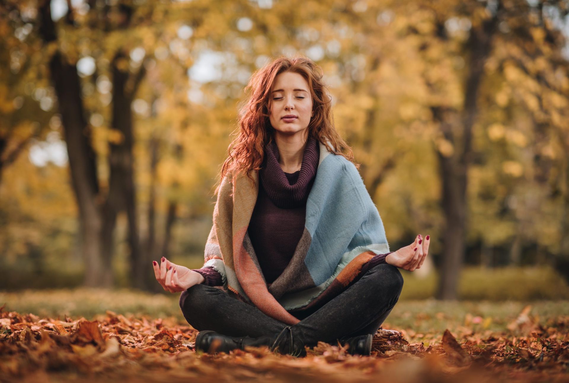 Woman with red hair meditating in lotus position in a park surrounded by autumn leaves.