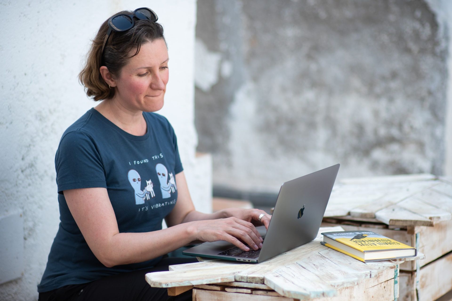 Woman typing on a laptop, outdoors. She wears a blue t-shirt with alien art. Book and sunglasses on a wooden bench.