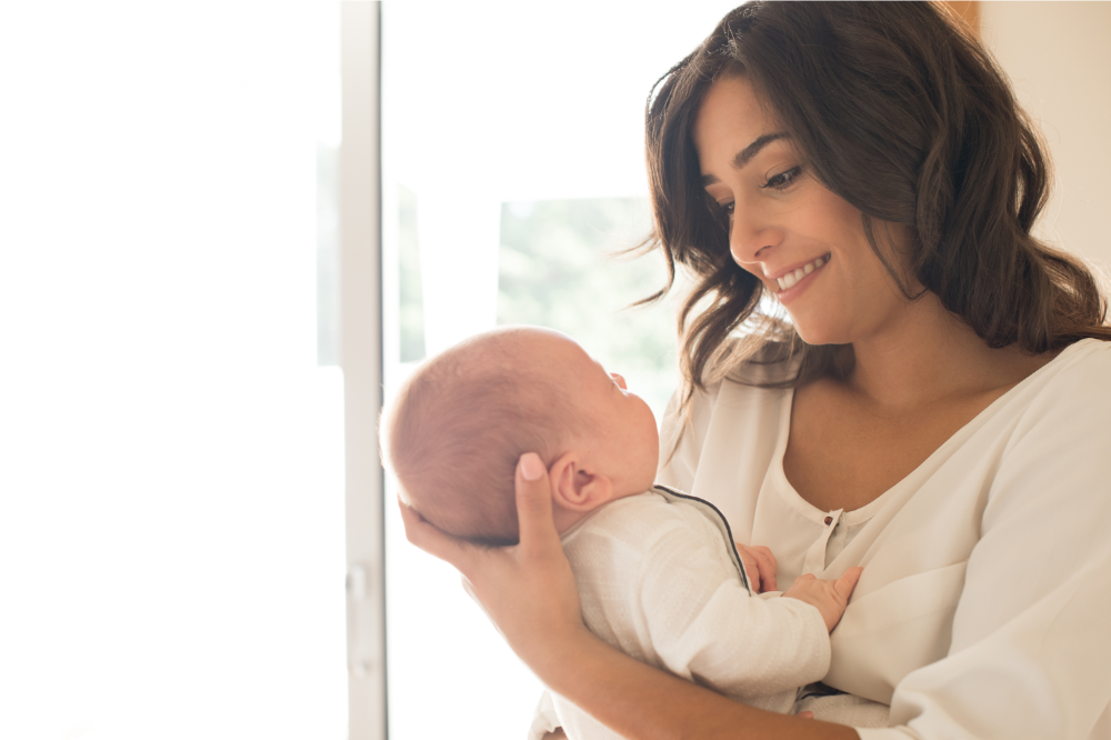Woman smiles, holding a baby near a bright window.