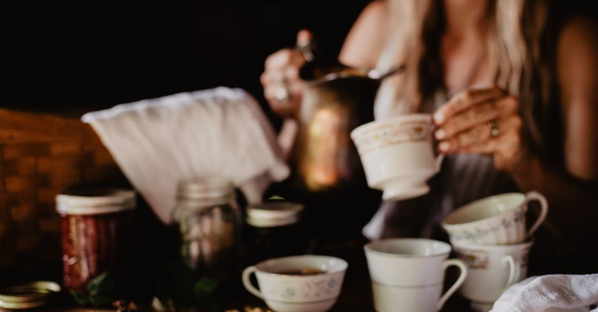 Woman pouring tea from a golden pot into teacups on a table, jars and cloth nearby.