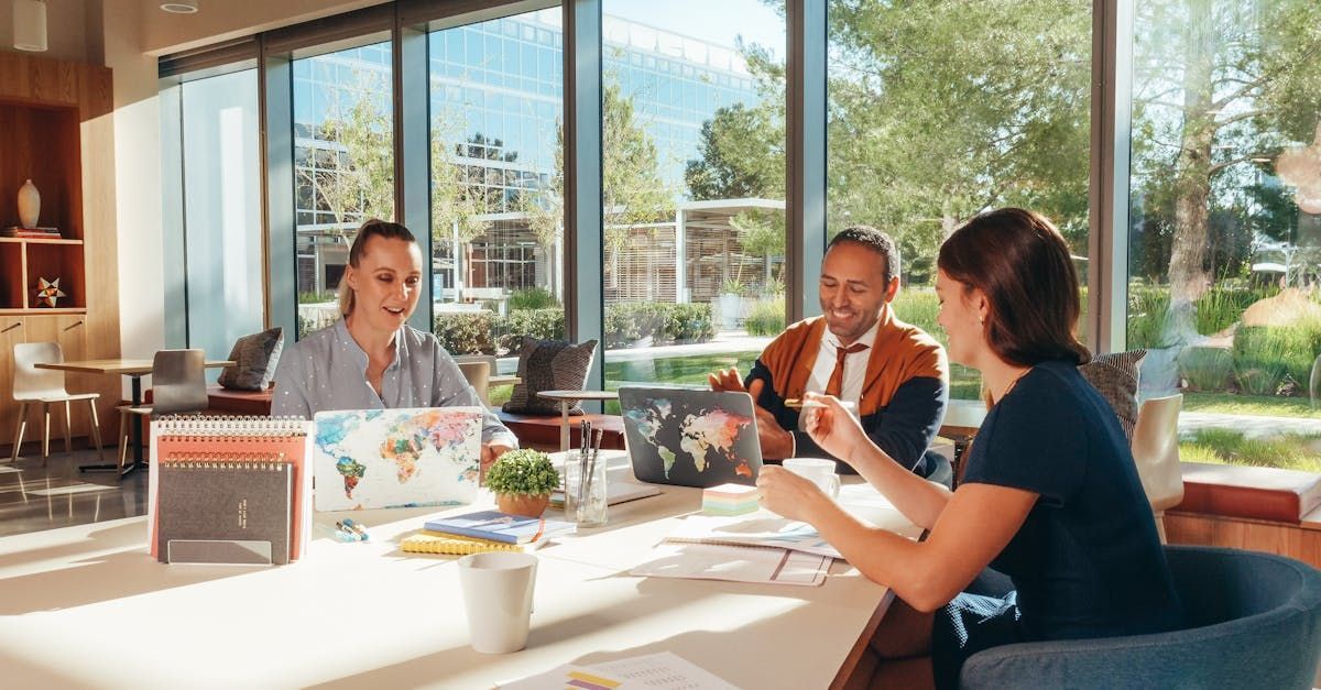Three people collaborating at a table with laptops, world maps, in a bright modern office.
