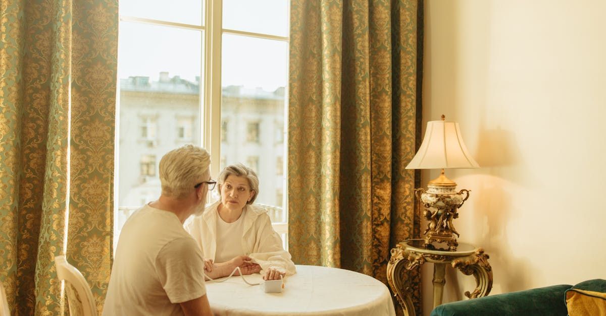 Elderly couple at a round table in a room with ornate curtains, a lamp, and a window.