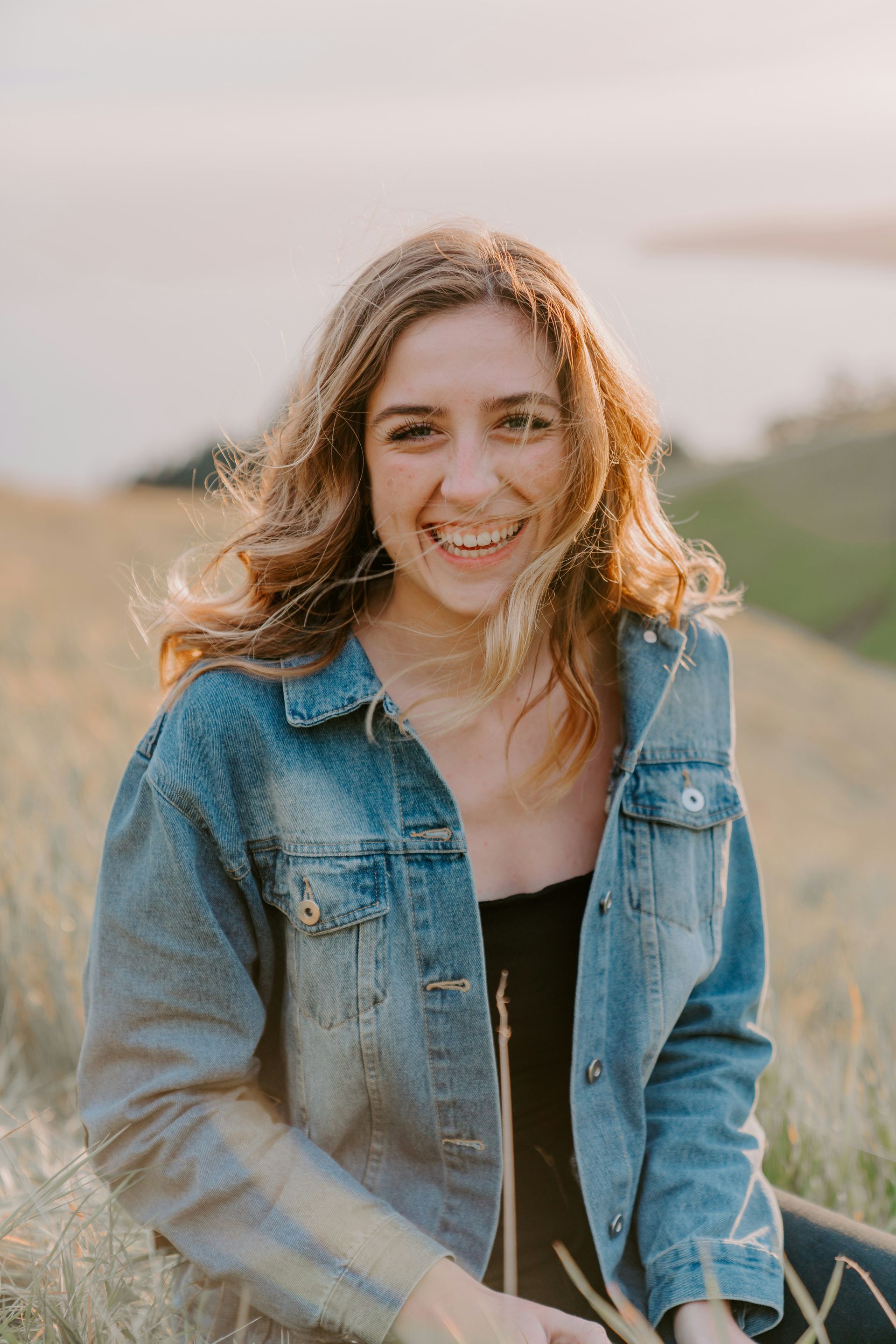 A smiling person with wavy hair wears a denim jacket, seated in a grassy, sunlit field overlooking a hillside.