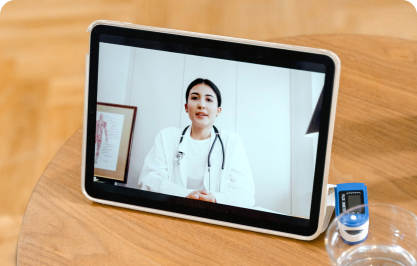 A doctor in a telehealth session, sitting on a wooden table with an oximeter and glass of water.