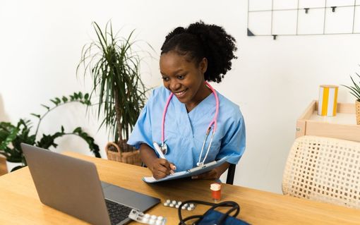 Woman in blue scrubs with a stethoscope smiles while taking notes at a desk with laptop and medication.
