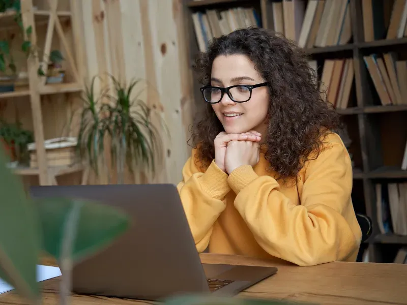 Woman with curly hair wearing glasses, looking at laptop screen. Leaning on hands, smiling; bookshelves in background.