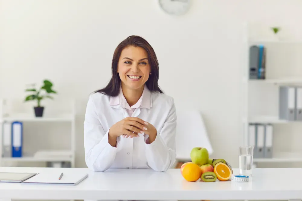 Smiling nutritionist in white coat at desk with fruit and water.