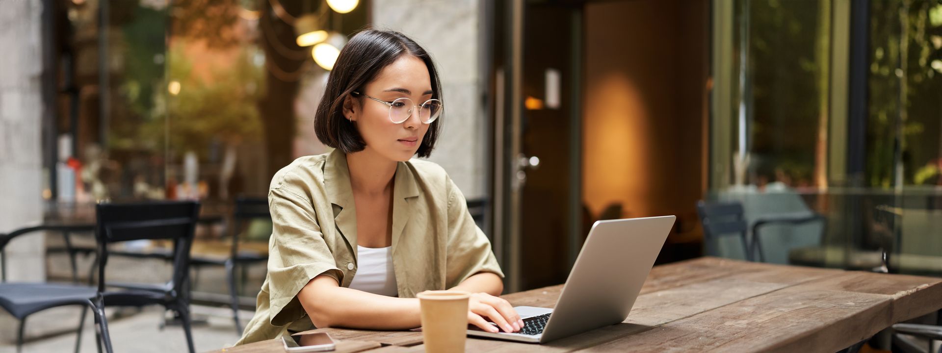 Woman working on a laptop at a cafe table. She wears glasses and a tan jacket.