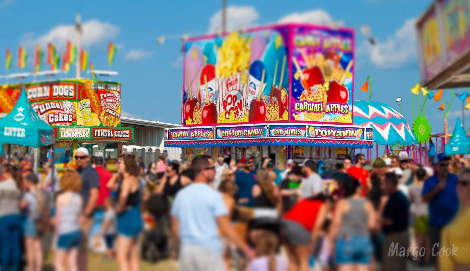 Crowded carnival midway with colorful food booths, bright banners, and people walking under a blue sky