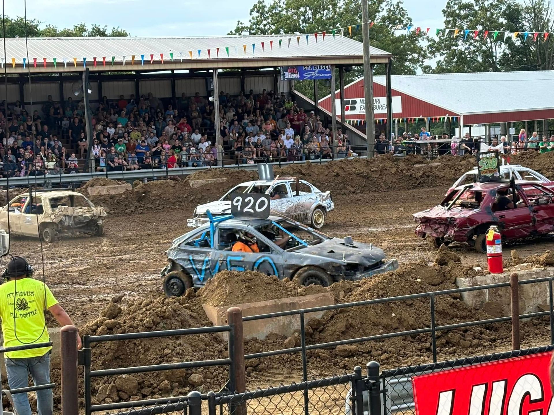 Demolition derby cars crash in a muddy arena with a crowded grandstand behind them.