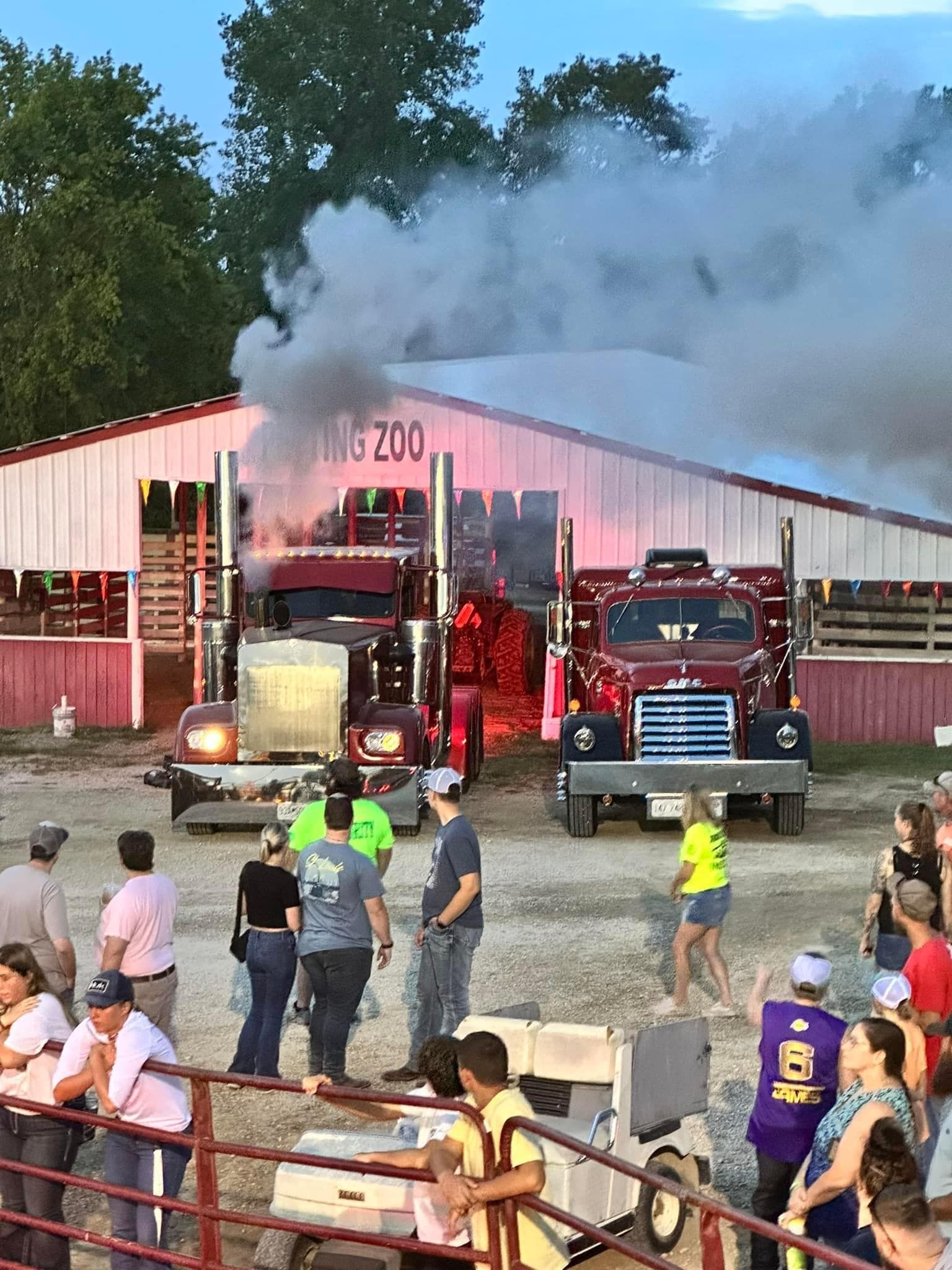 Fire trucks outside a smoke-filled red barn as people watch from a fence line