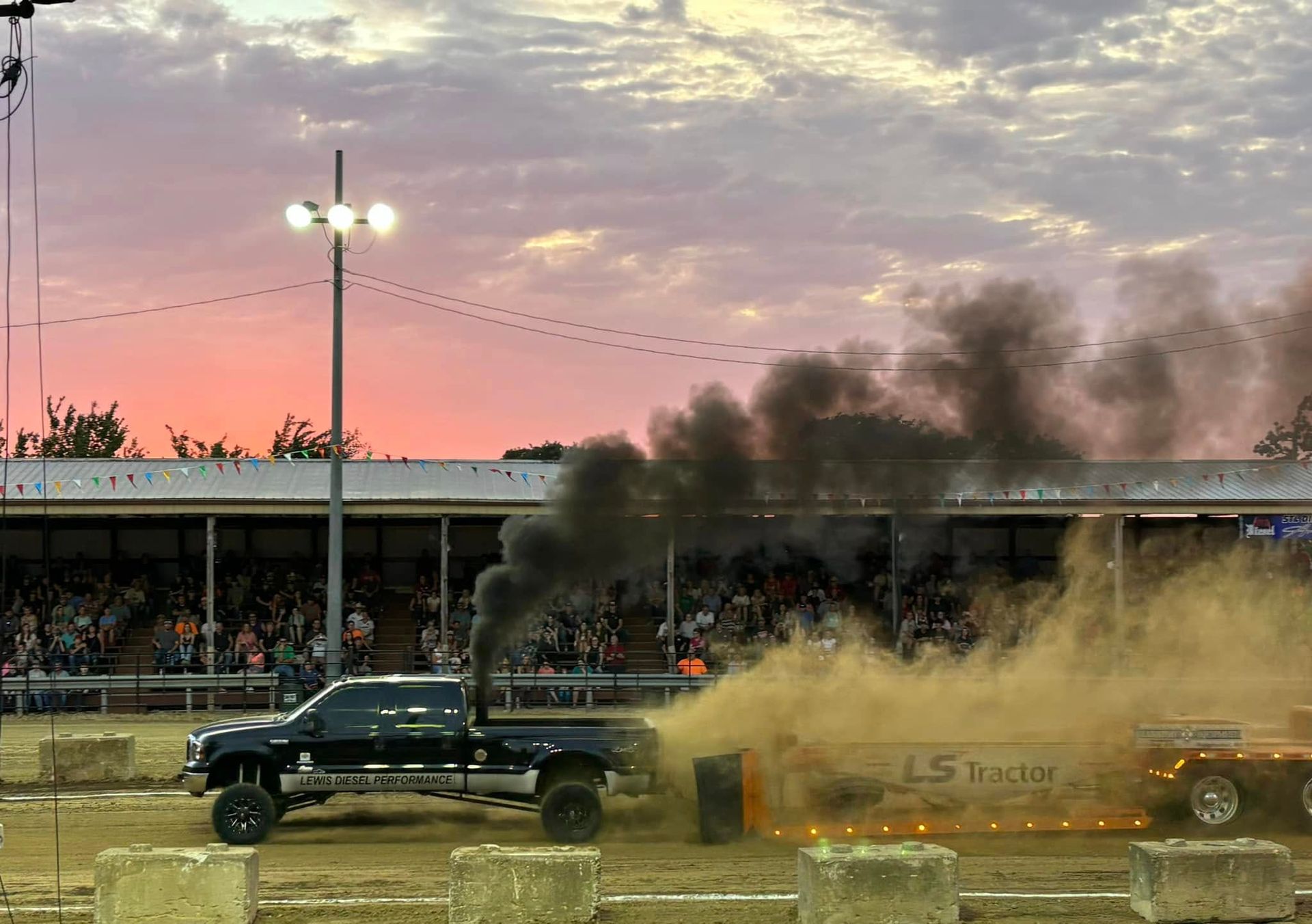 Black pickup truck pulling a smoking sled at a dirt-track event at sunset