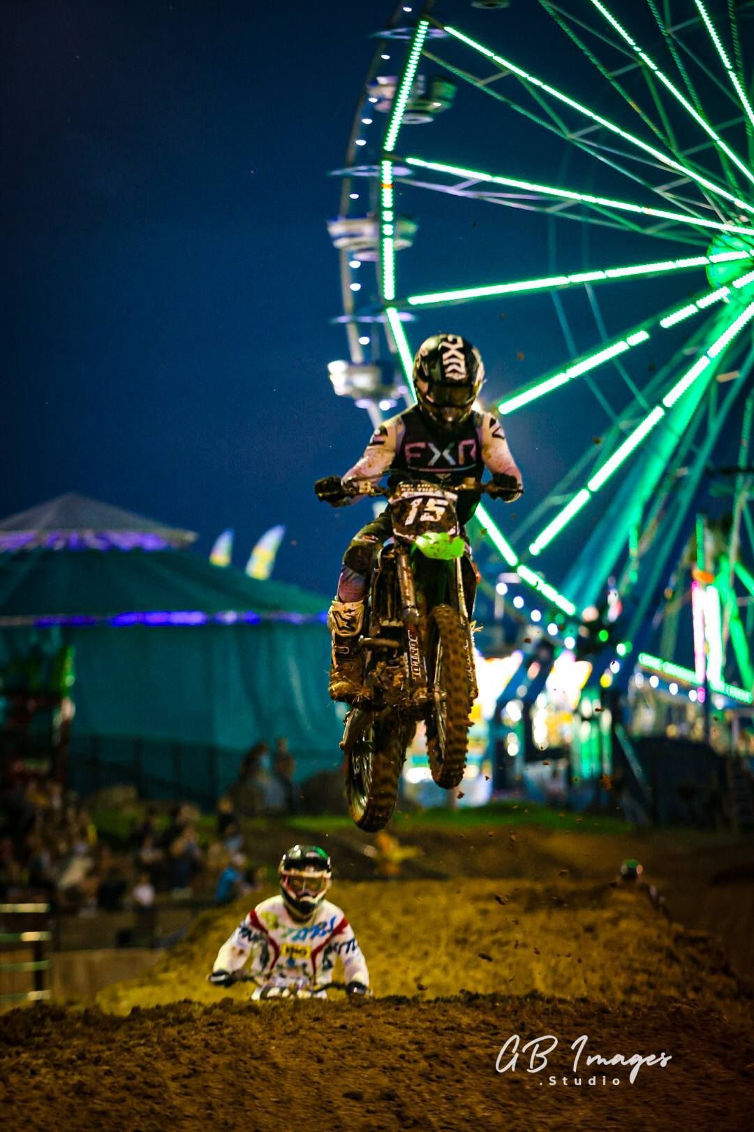 Motocross rider airborne at night near a brightly lit Ferris wheel