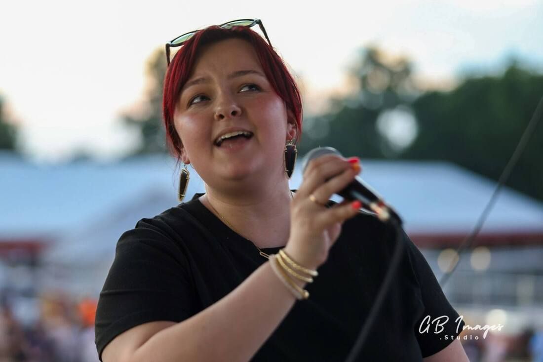 Singer holding a microphone on an outdoor stage, wearing a black shirt and red headband.