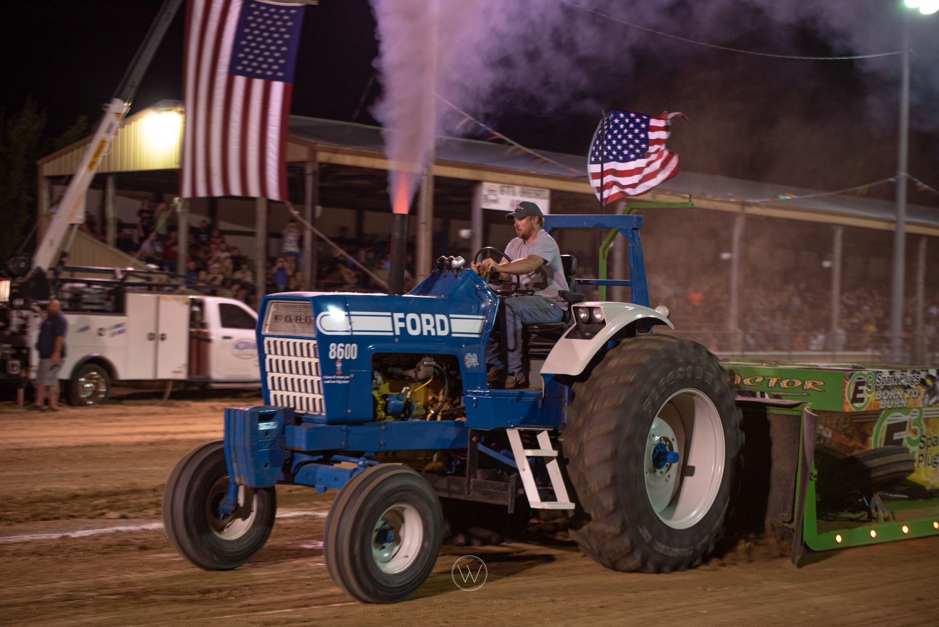 Blue Ford tractor pulling a sled at a nighttime dirt track, with smoke and American flags waving.