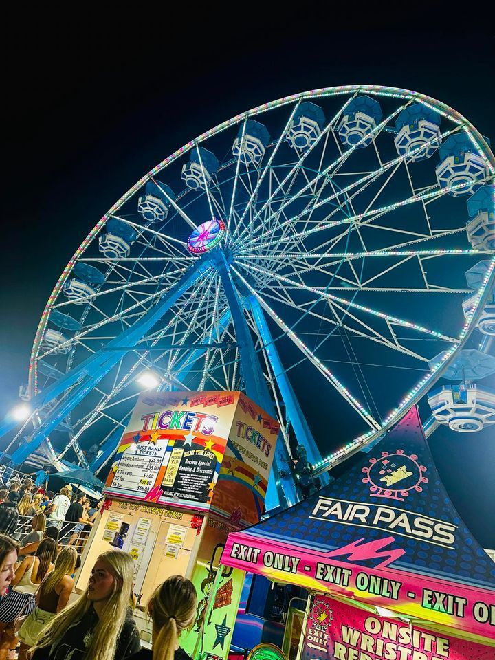 Nighttime Ferris wheel at a brightly lit amusement park booth with colorful signs and people below