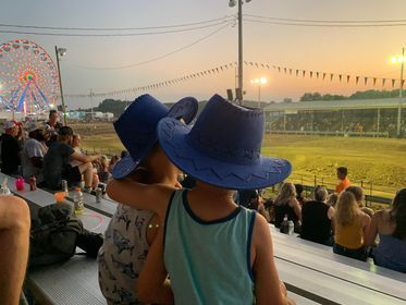 Two children in blue cowboy hats watch a sunset event from bleachers near a lit field and Ferris wheel