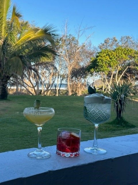 Three glasses of drinks are sitting on a balcony overlooking a lush green field.