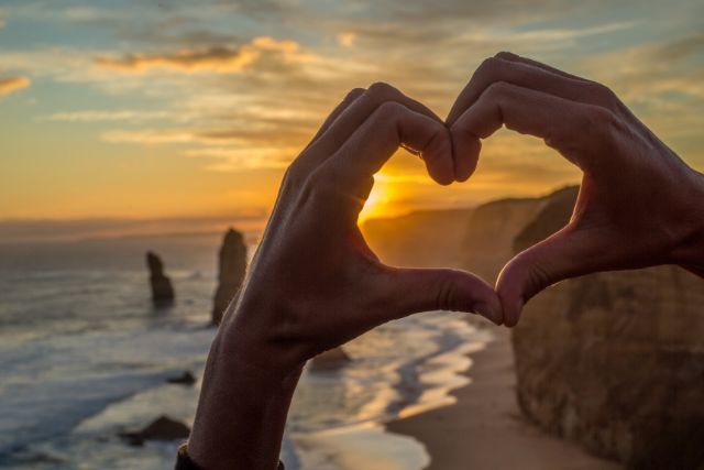 A person is making a heart shape with their hands at sunset.