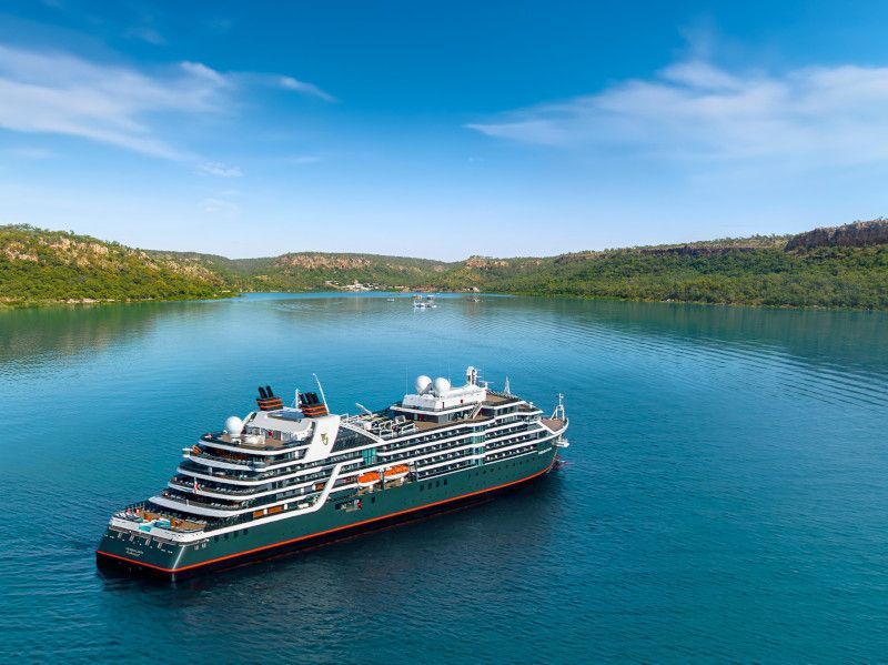 Cruise ship on calm blue water, surrounded by lush green hills under a bright blue sky.