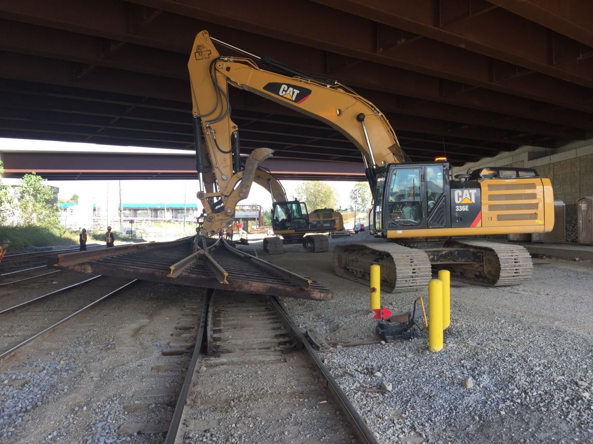 A large yellow excavator is working on train tracks under a bridge.