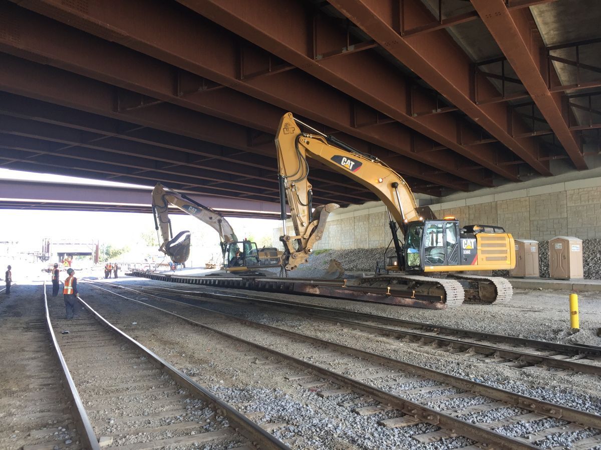 Two excavators are working on train tracks under a bridge.