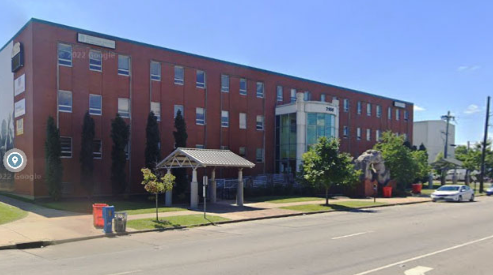 Brick building with several windows, an awning, and a glass-enclosed section. Trees and parked cars are visible.
