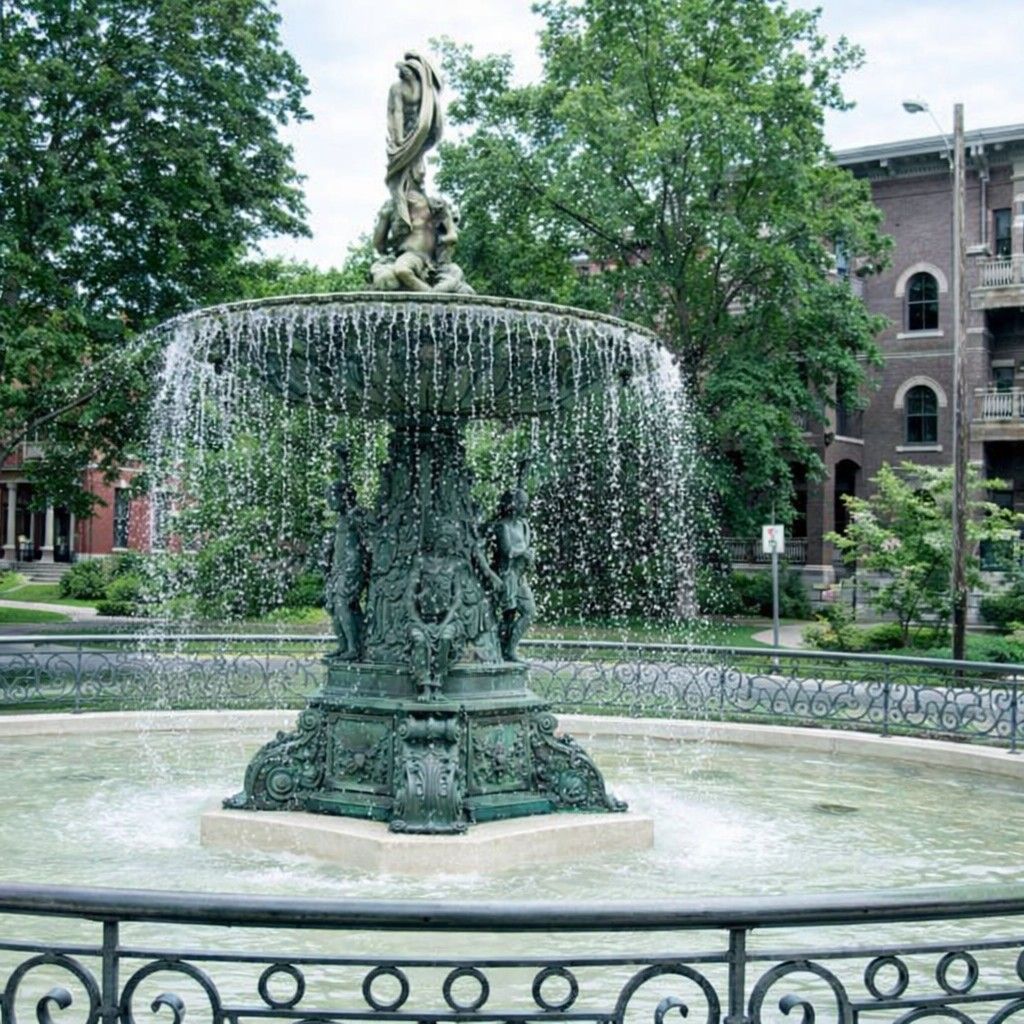 A large ornate bronze fountain with water cascading into a pool, surrounded by a wrought-iron fence and greenery.