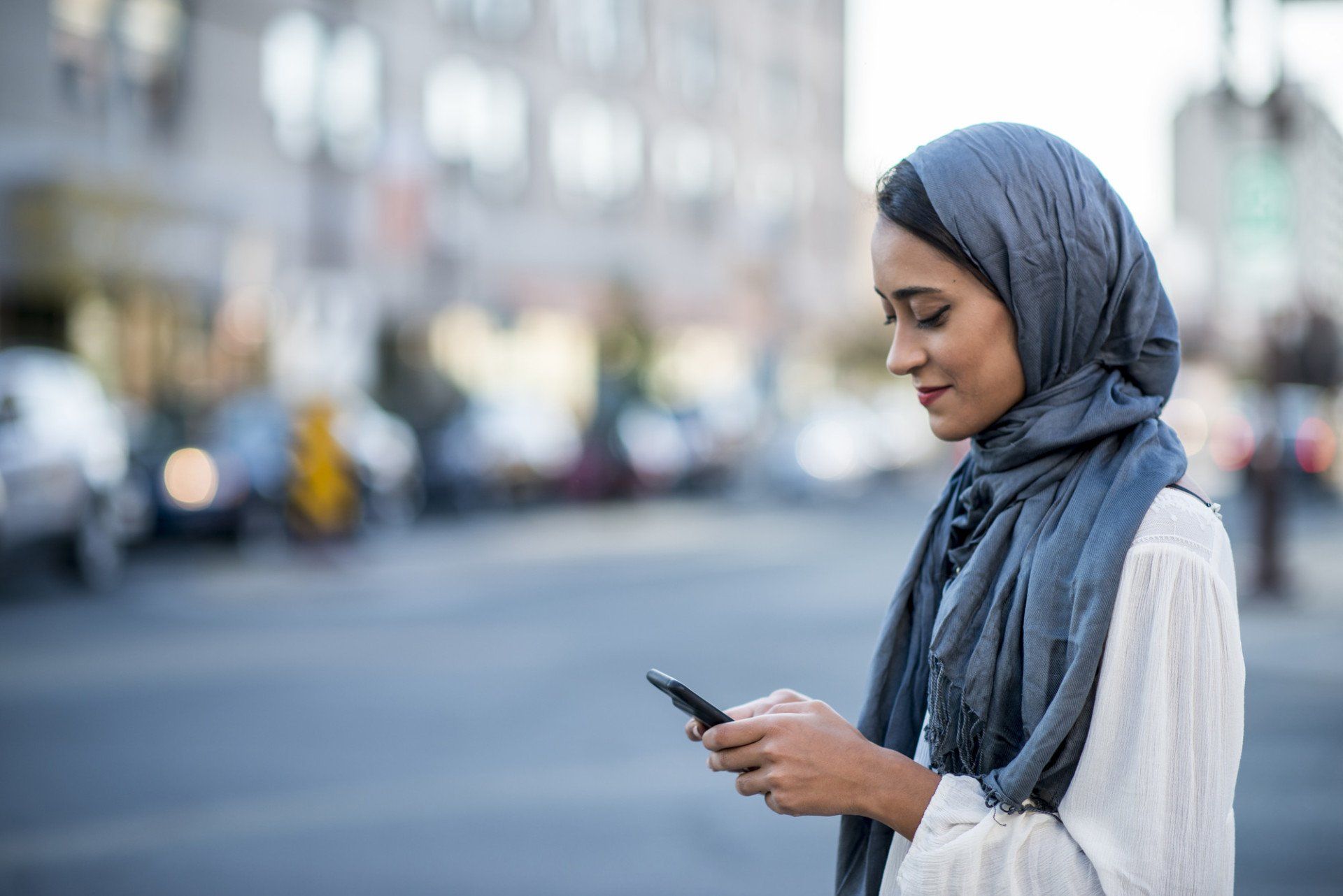 A Muslim woman is outdoors on a sunny day. She is wearing casual clothing and a head scarf. She is standing near a road and sending a message with her smartphone.