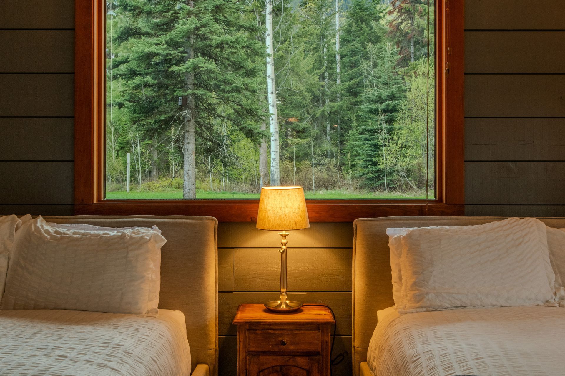 A bedroom with two beds , a nightstand , a lamp and a large window.