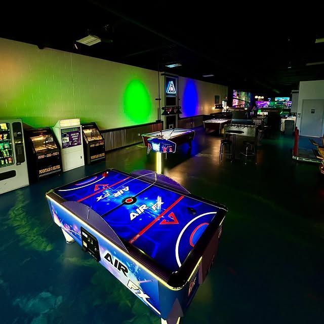A dark indoor arcade with an air hockey table in the foreground, illuminated by green and blue wall lights.