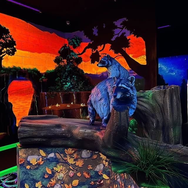 Two raccoon statues sit on a large log in a blacklight-illuminated indoor miniature golf course with sunset murals.