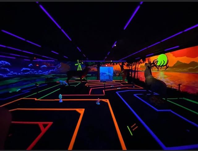 A glow-in-the-dark indoor miniature golf course with neon-lit paths, animal statues, and a sunset mural backdrop.