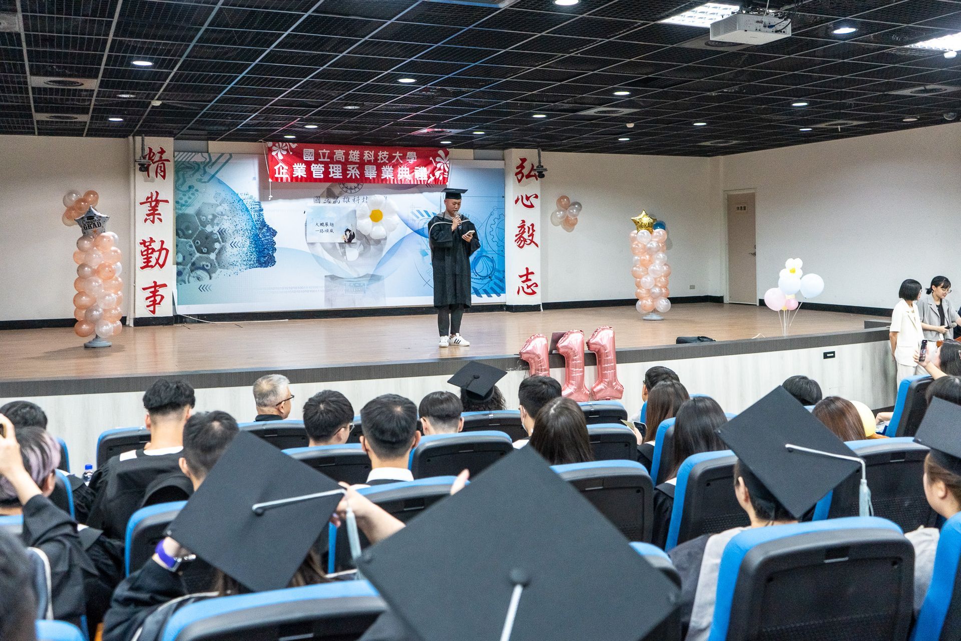 一名男子站在舞台上，前方是一群戴著畢業帽的人。