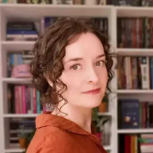 A woman with curly hair is standing in front of a bookshelf.