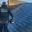 A man wearing a felt that roofing vest is standing on a roof holding a chainsaw.