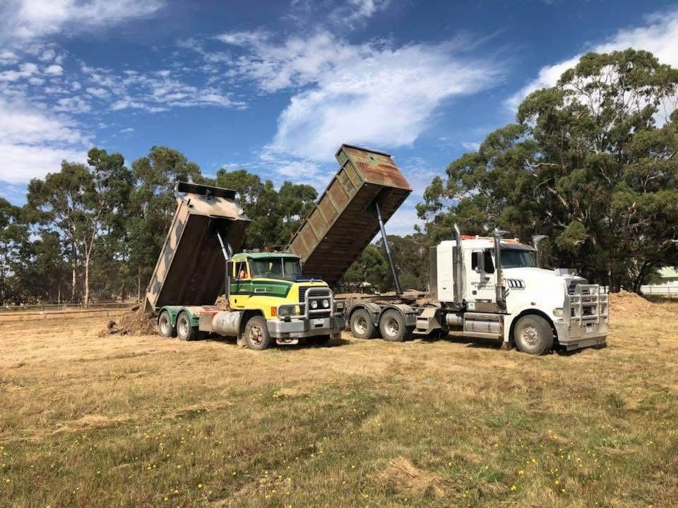 Two dump trucks unloading dirt in a field, one green, one white, under a blue sky — Beaufort & District Earthworks in Beaufort, VIC