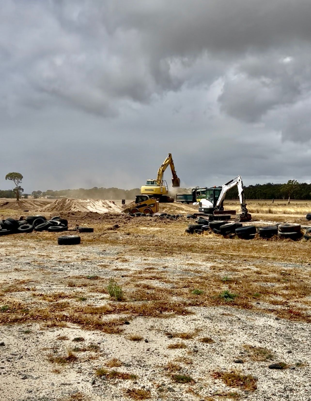 Two excavators working on a barren field under a cloudy sky. Scattered tires and dirt piles — Beaufort & District Earthworks in Beaufort, VIC